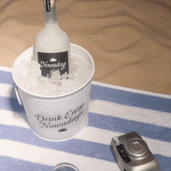 Nowadays High Dose THC infused bottle chilling in an ice bucket reading Drink Easy on a striped beach towel, lifestyle shot