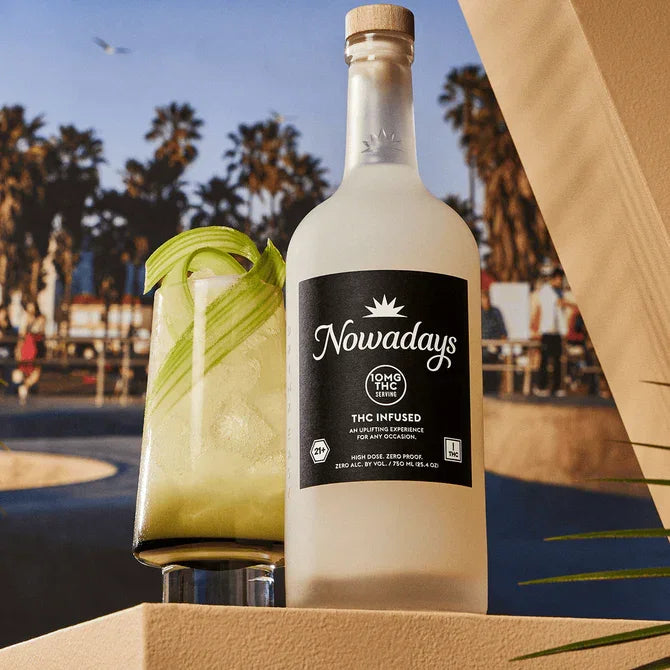 Nowadays High Dose 166mg THC infused 750ml bottle beside a garnished cocktail with palm trees behind, outdoor lifestyle